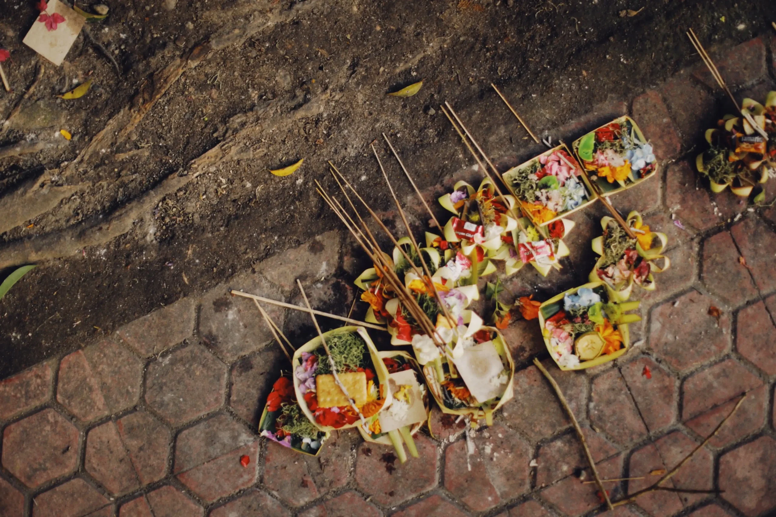 Aerial view of multiple canang sari — traditional Balinese Hindu offerings made from woven palm leaf baskets filled with colorful flowers, rice, crackers, and incense sticks — arranged on stone pavement