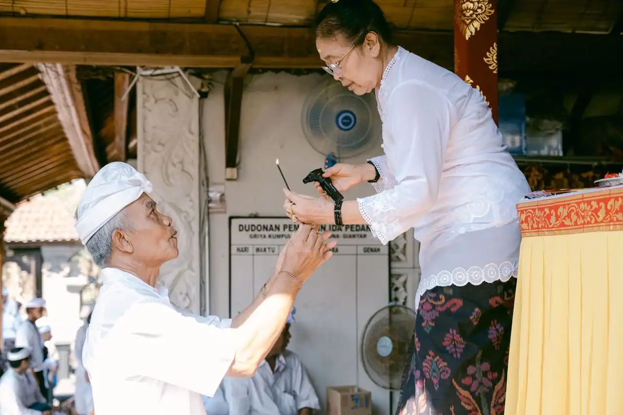 An elderly Balinese man in white ceremonial attire and udeng headband receiving a lit incense stick from a woman in a white kebaya and traditional batik kain skirt during a Hindu temple ceremony in Bali
