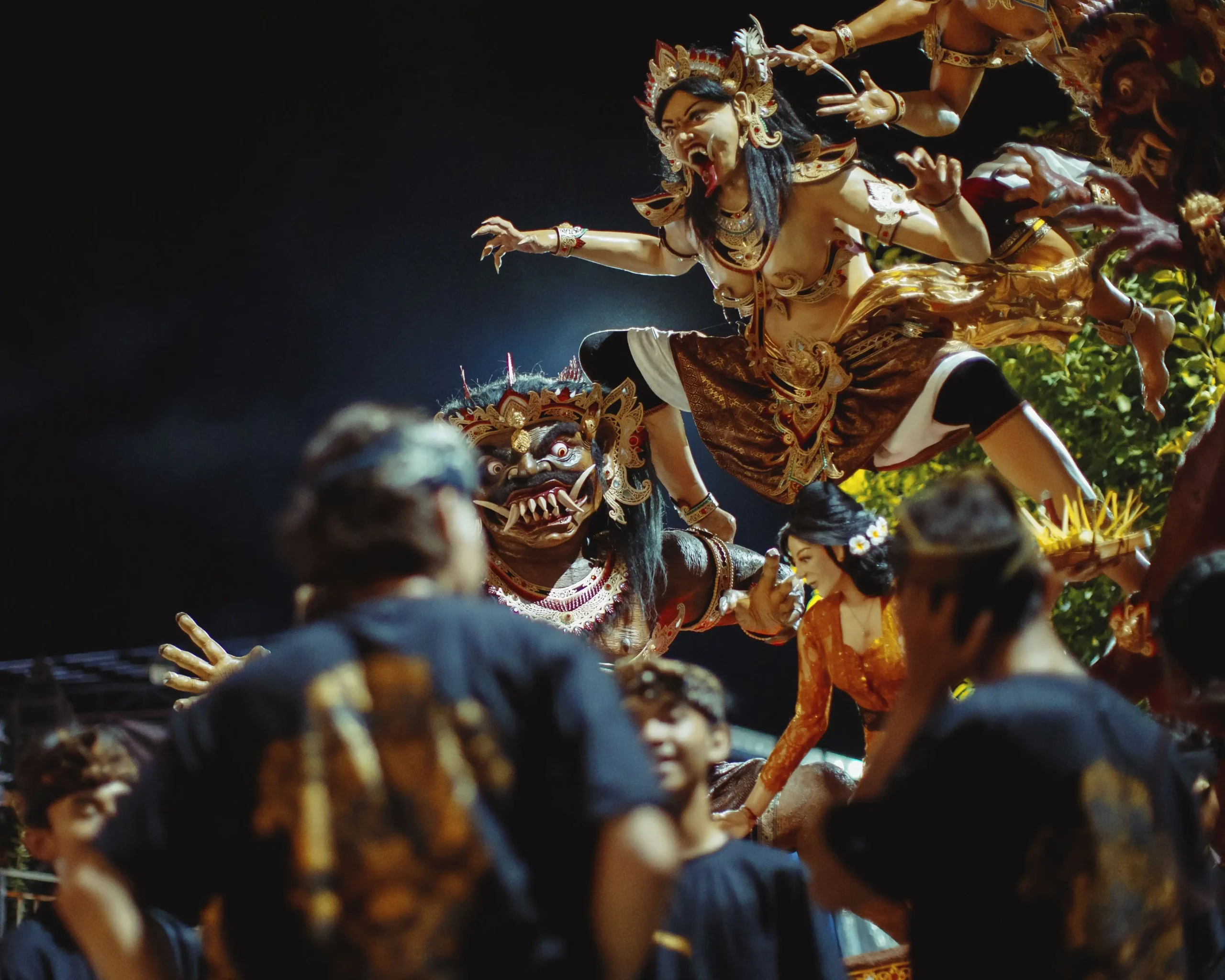 A towering ogoh-ogoh demon effigy with elaborate gold and dark costumes being carried through a crowd during the Balinese Ngrupuk parade on the eve of Nyepi, the Balinese Day of Silence, with participants in traditional attire surrounding it against a dark night sky