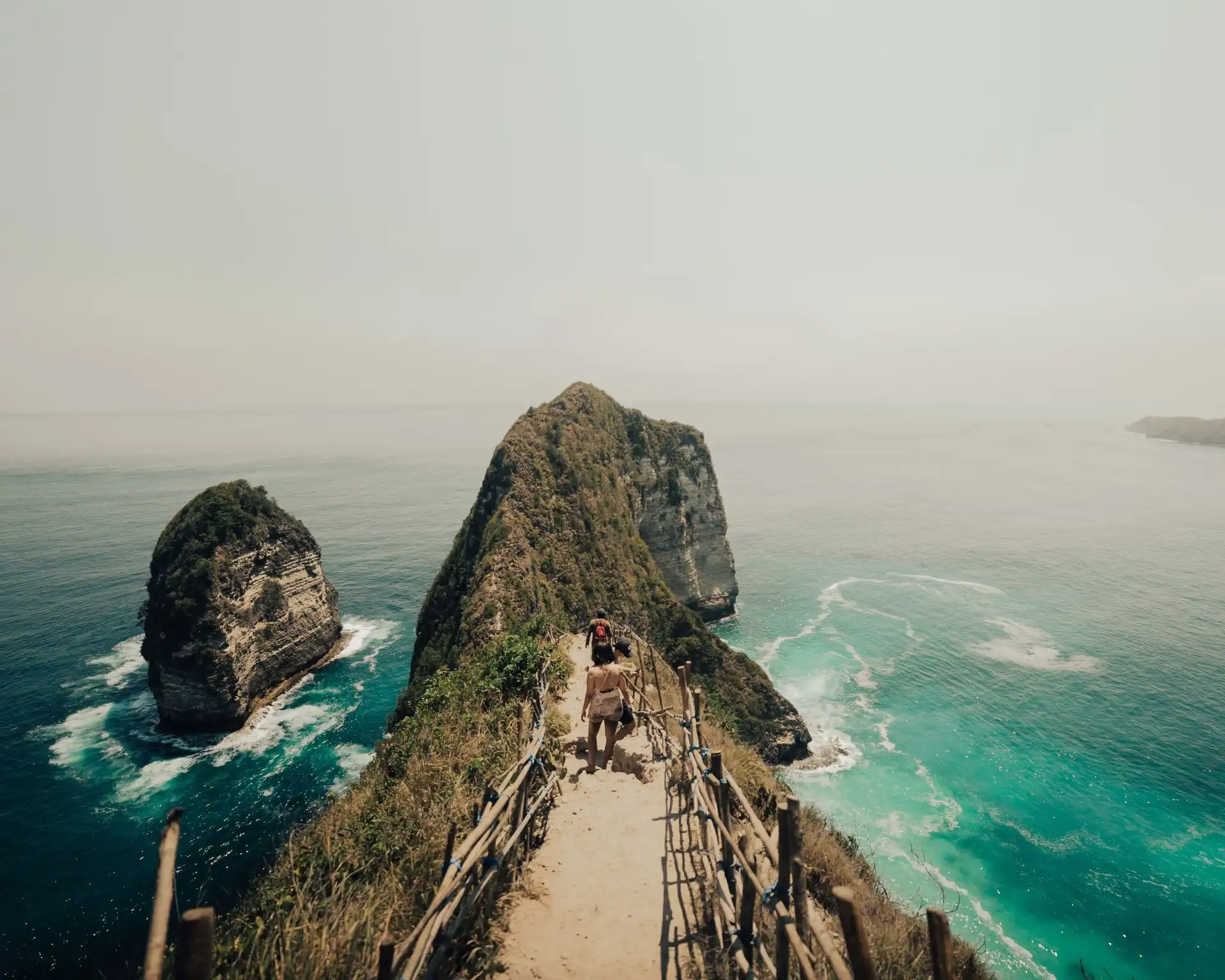 Two hikers walking along a narrow cliff trail with a wooden railing on Nusa Penida, Bali, with dramatic limestone rock formations rising from turquoise ocean on both sides under a hazy sky