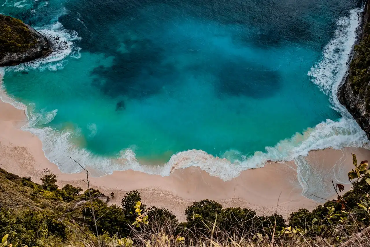 Aerial overhead view of a secluded crescent beach on Nusa Penida with vivid turquoise and teal water gradients, white foamy waves, and lush cliff vegetation framing the shoreline
