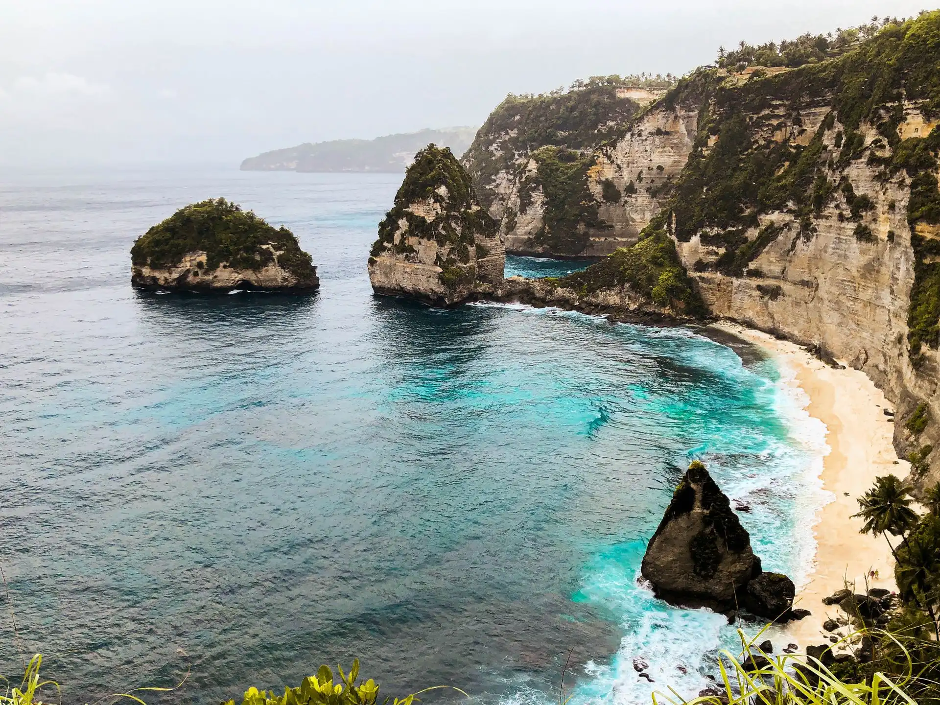 Elevated coastal view of a secluded beach on Nusa Penida flanked by towering white limestone cliffs and multiple rock formations rising from turquoise water, with a lone palm tree and tiny figures visible on the sandy shoreline