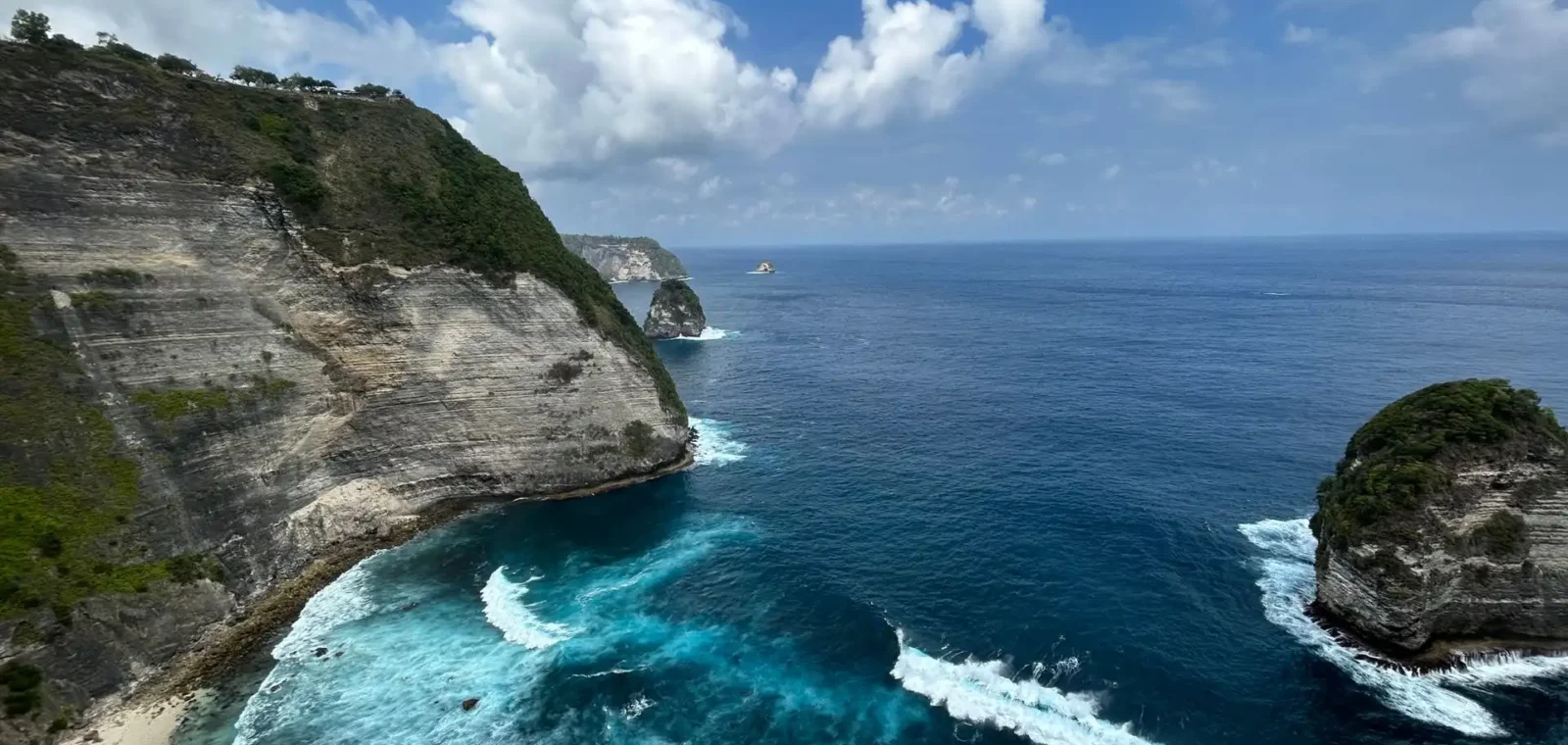 Panoramic coastal view from a clifftop on Nusa Penida, Bali, with towering stratified white limestone cliffs, crashing turquoise waves, a small rocky islet, and a deep blue ocean stretching to the horizon under a bright cloudy sky