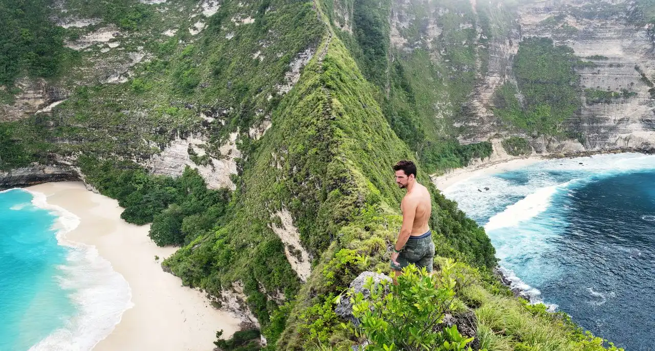 A shirtless man standing on a lush green clifftop viewpoint on Nusa Penida, Bali, overlooking two secluded white sand beaches with vivid turquoise waves on either side of a dramatic green ridge