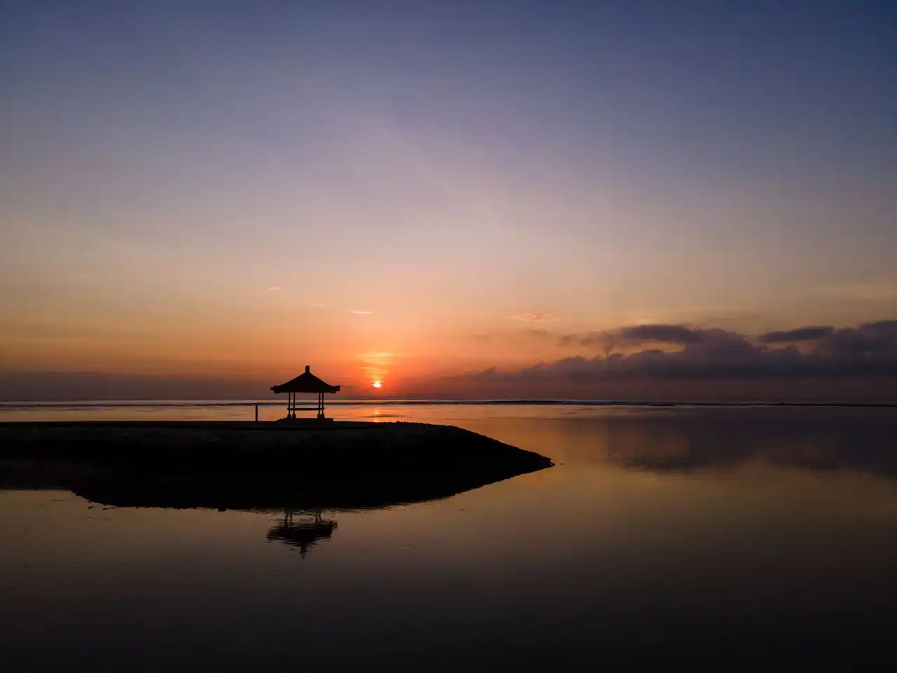 Silhouette of a traditional Balinese pavilion on a narrow coastal strip at sunrise during Nyepi, with a vivid orange and purple sky reflected on the calm, still water surrounding it