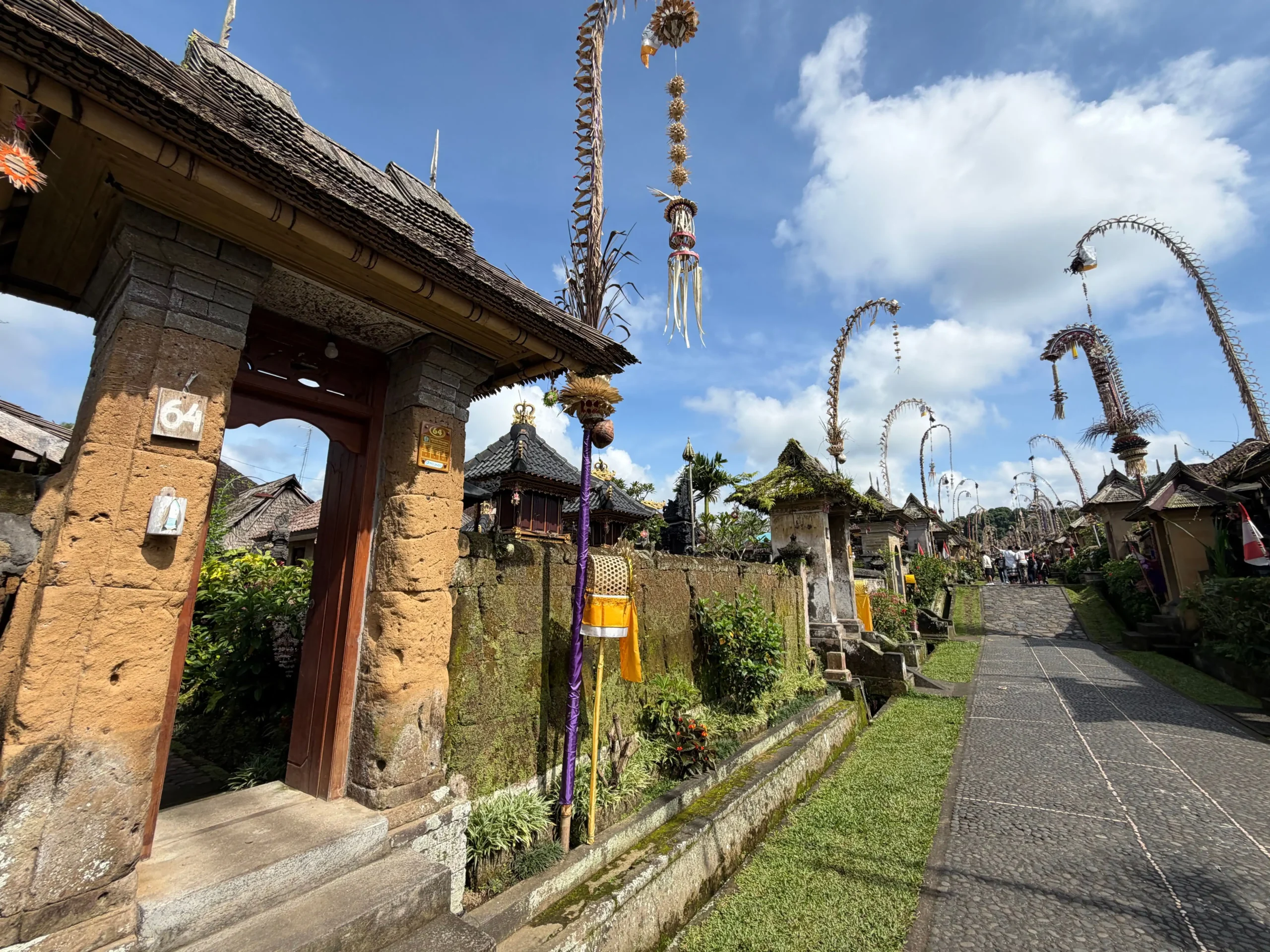 A traditional street in Penglipuran Village, Bali, lined with moss-covered stone compound walls and wooden entry gates, decorated with tall curved penjor poles adorned with woven bamboo and hanging ornaments under a blue sky