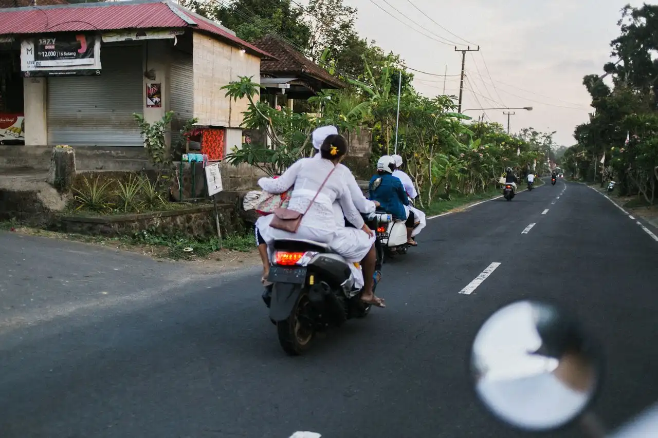 Balinese locals dressed in white ceremonial kebaya and udeng riding motorbikes along a rural road in Bali, likely heading to or from a temple ceremony, with tropical vegetation and small roadside shops in the background