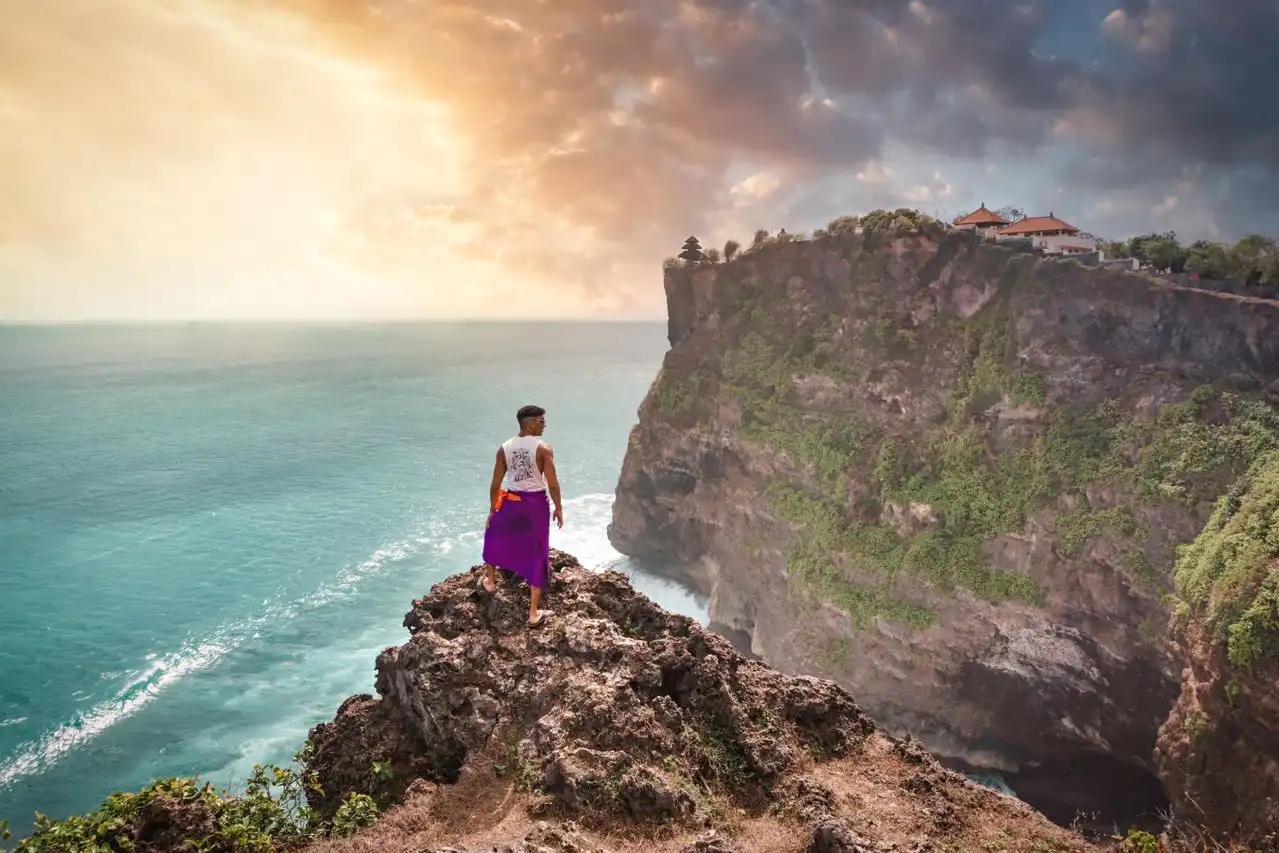 A person wearing a purple sarong stands on a rugged limestone outcrop overlooking the turquoise ocean at Uluwatu, with Pura Luhur Uluwatu temple visible on the clifftop to the right under a dramatic stormy sunset sky