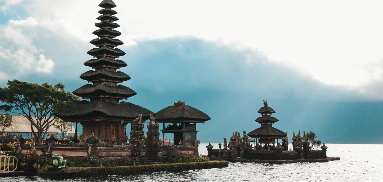 Pura Ulun Danu Bratan, a Balinese Hindu water temple featuring a tall multi-tiered meru tower and smaller shrines built on the edge of Lake Bratan, surrounded by misty mountains and dramatic clouds