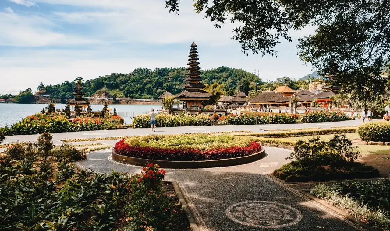 Landscaped gardens with colorful flower beds and a circular roundabout at Pura Ulun Danu Bratan, with the iconic multi-tiered meru tower, thatched-roof shrines, and Lake Bratan backed by forested hills visible in the background