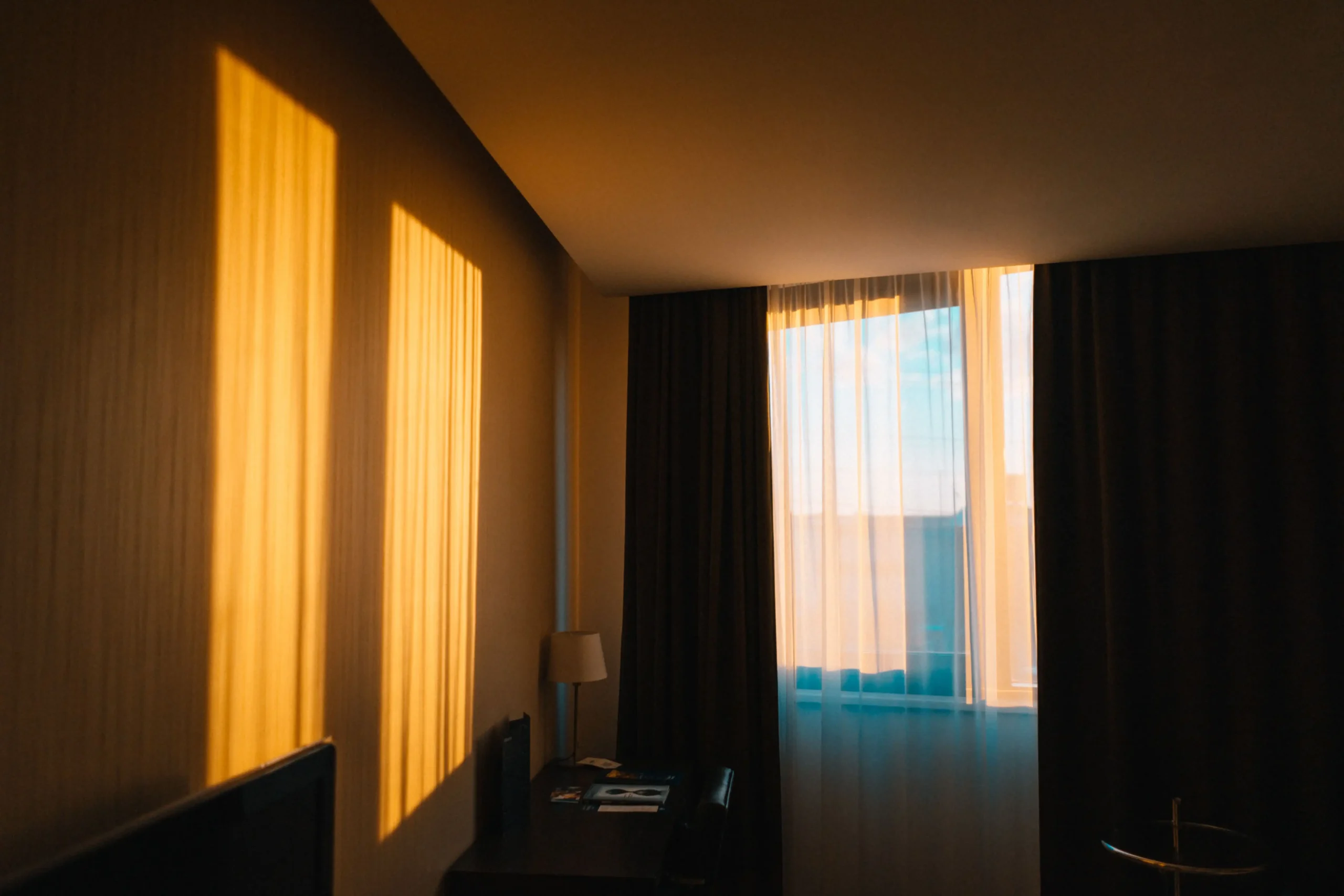 A dimly lit hotel room bathed in warm golden sunlight streaming through sheer curtains, casting rectangular light patterns on the wall, with a small desk lamp and blackout curtains visible in the background
