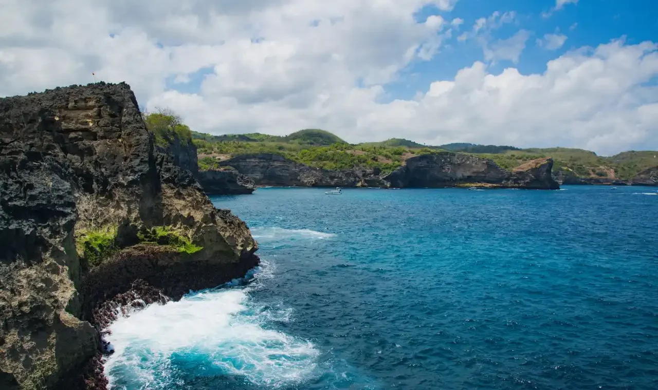 Rugged dark volcanic rock cliffs with crashing waves on the coastline of Nusa Penida, Bali, with a small boat visible on the deep blue ocean and green rolling hills stretching along the coast in the background