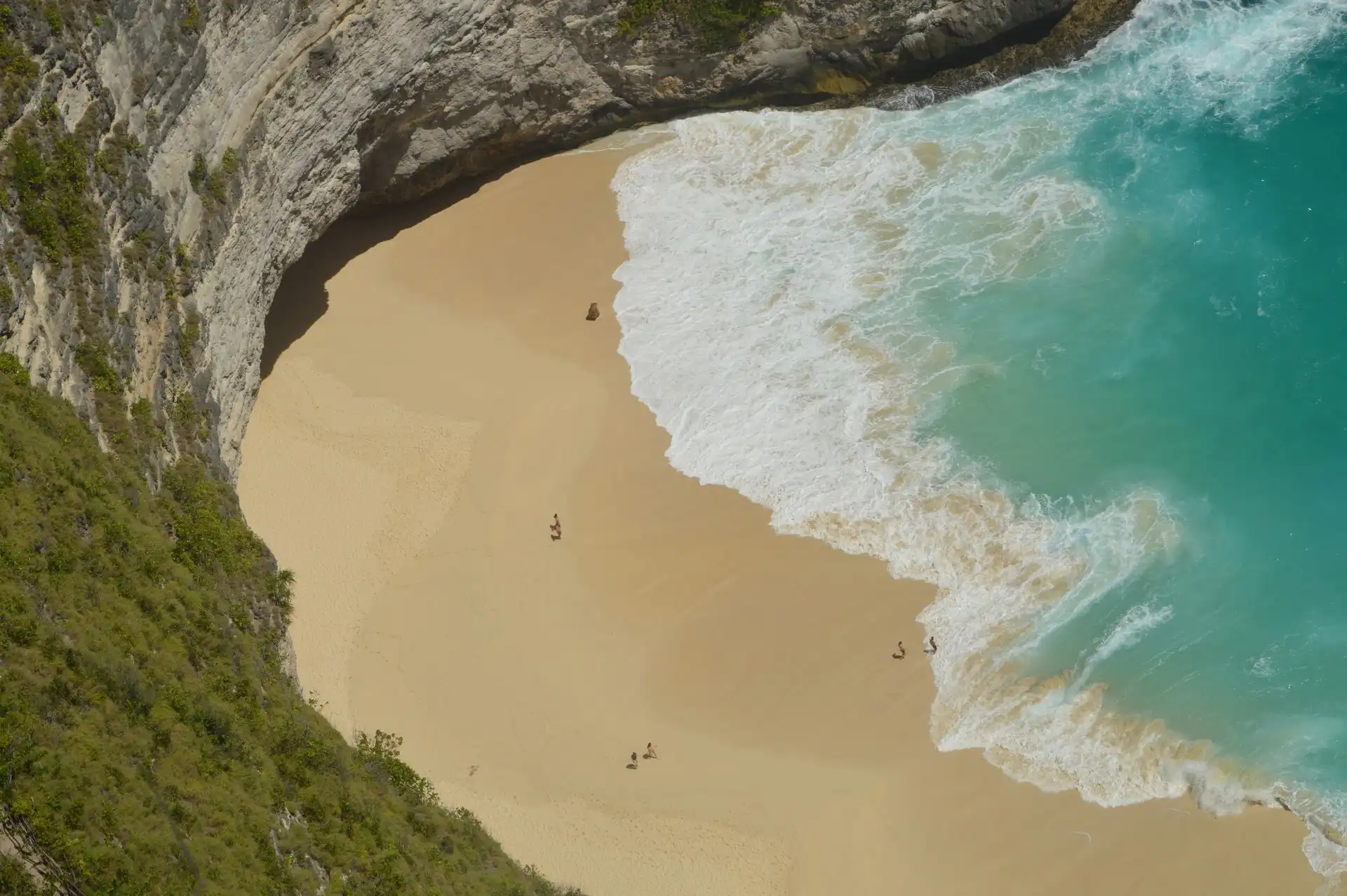 Aerial view of a curved secluded white sand beach tucked beneath towering white limestone cliffs on Nusa Penida, with turquoise waves rolling in and a handful of tiny visitors visible on the empty shoreline