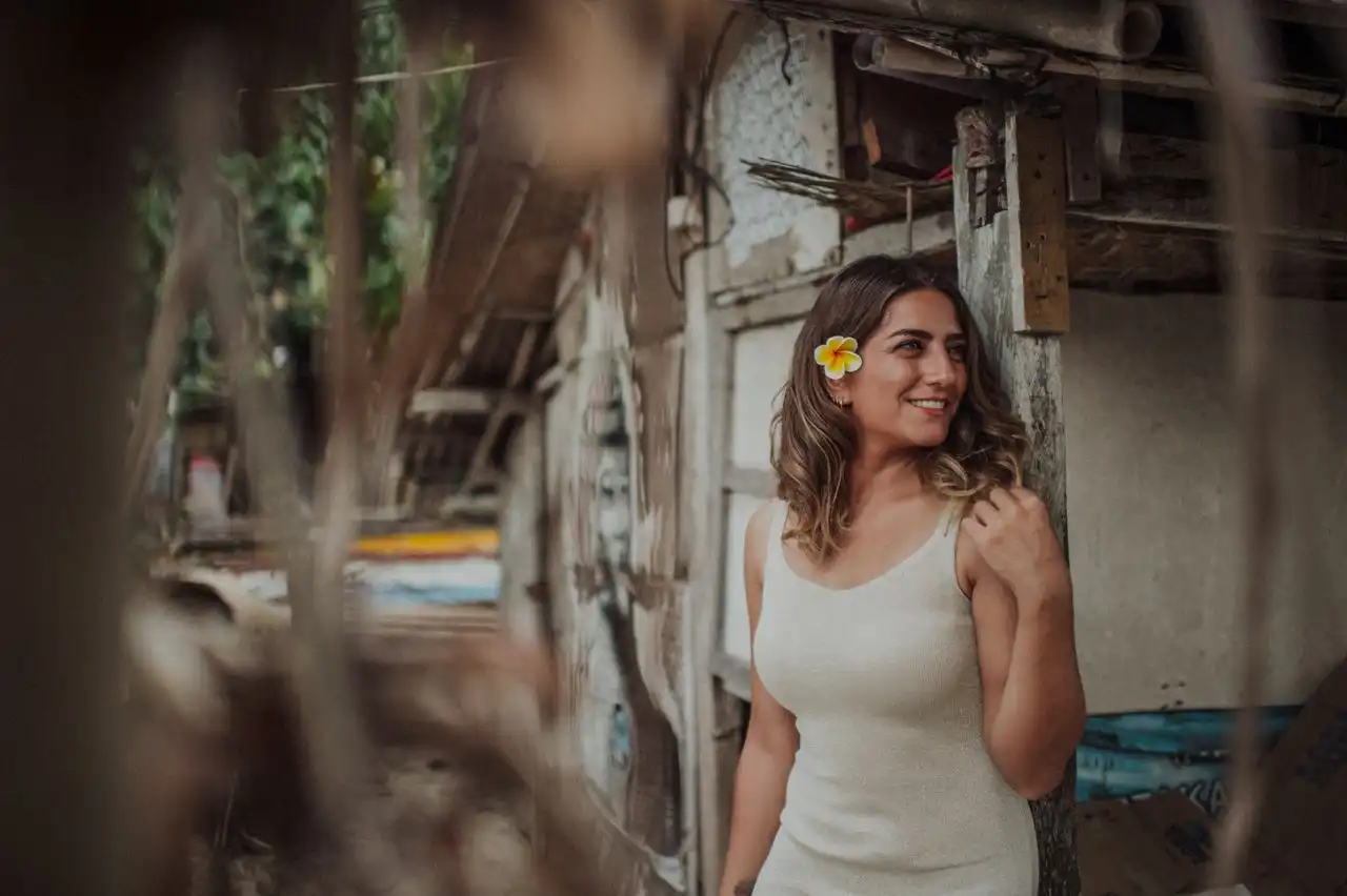 A smiling woman wearing a white dress and a frangipani flower in her hair, leaning against a rustic wooden doorway in Bali, photographed through a blurred foreground with a bokeh background