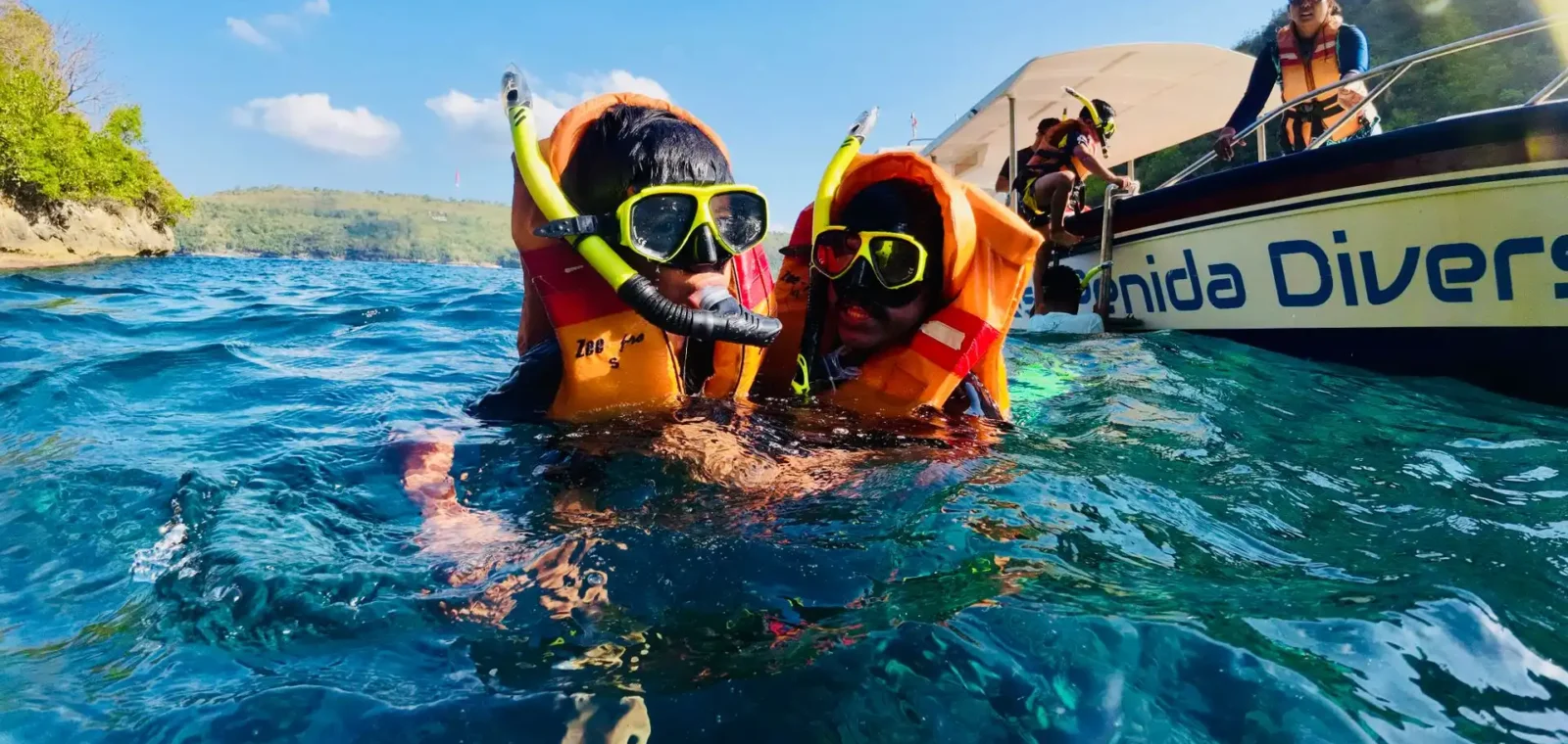 Two snorkelers in orange life jackets and yellow dive masks floating in clear blue water off Nusa Penida, Bali, with a dive boat and other participants visible in the background