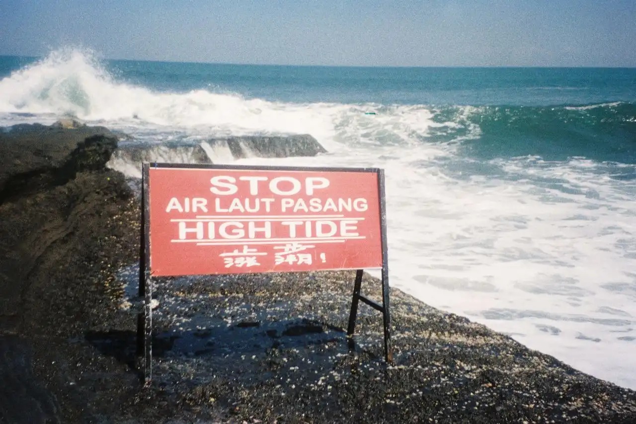 A red multilingual warning sign reading 'Stop – Air Laut Pasang – High Tide' in English, Indonesian, and Chinese, placed on a rocky coastal shoreline in Bali with powerful crashing waves in the background