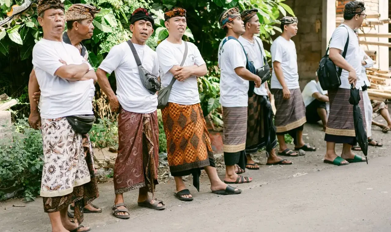 A group of Balinese men standing in a row along a street wearing traditional udeng headbands and colorful batik and endek sarongs with white t-shirts, gathered for a community ceremony or gotong royong event in Bali