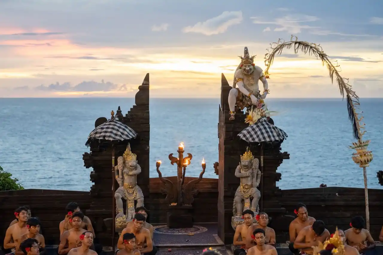 Kecak fire dance performance at Uluwatu Temple, Bali, with a performer in white Hanuman costume perched on a split stone candi bentar gate, flanked by black-and-white poleng umbrellas and guardian statues, while a chorus of bare-chested male dancers sit below against a dramatic ocean sunset backdrop