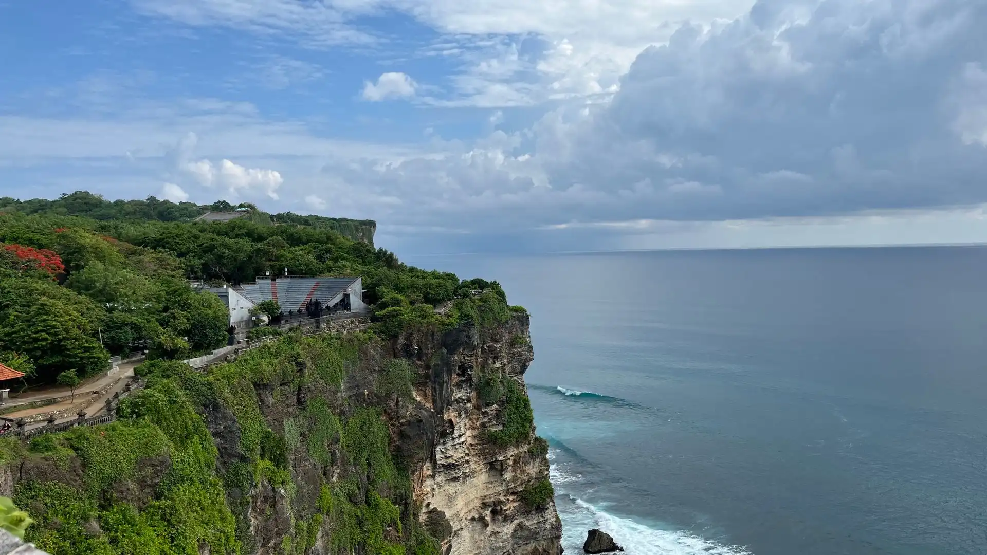 Dramatic clifftop view of Uluwatu Temple area in Bali, with lush green vegetation and temple structures perched on the edge of a steep limestone cliff overlooking the turquoise Indian Ocean below
