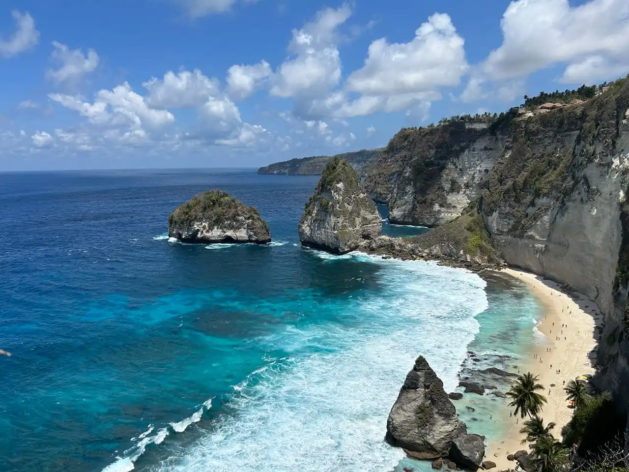 Aerial coastal view of a secluded white sand beach tucked beneath towering white limestone cliffs on Nusa Penida, with two large rock formations rising from vivid turquoise water and tiny beachgoers visible below