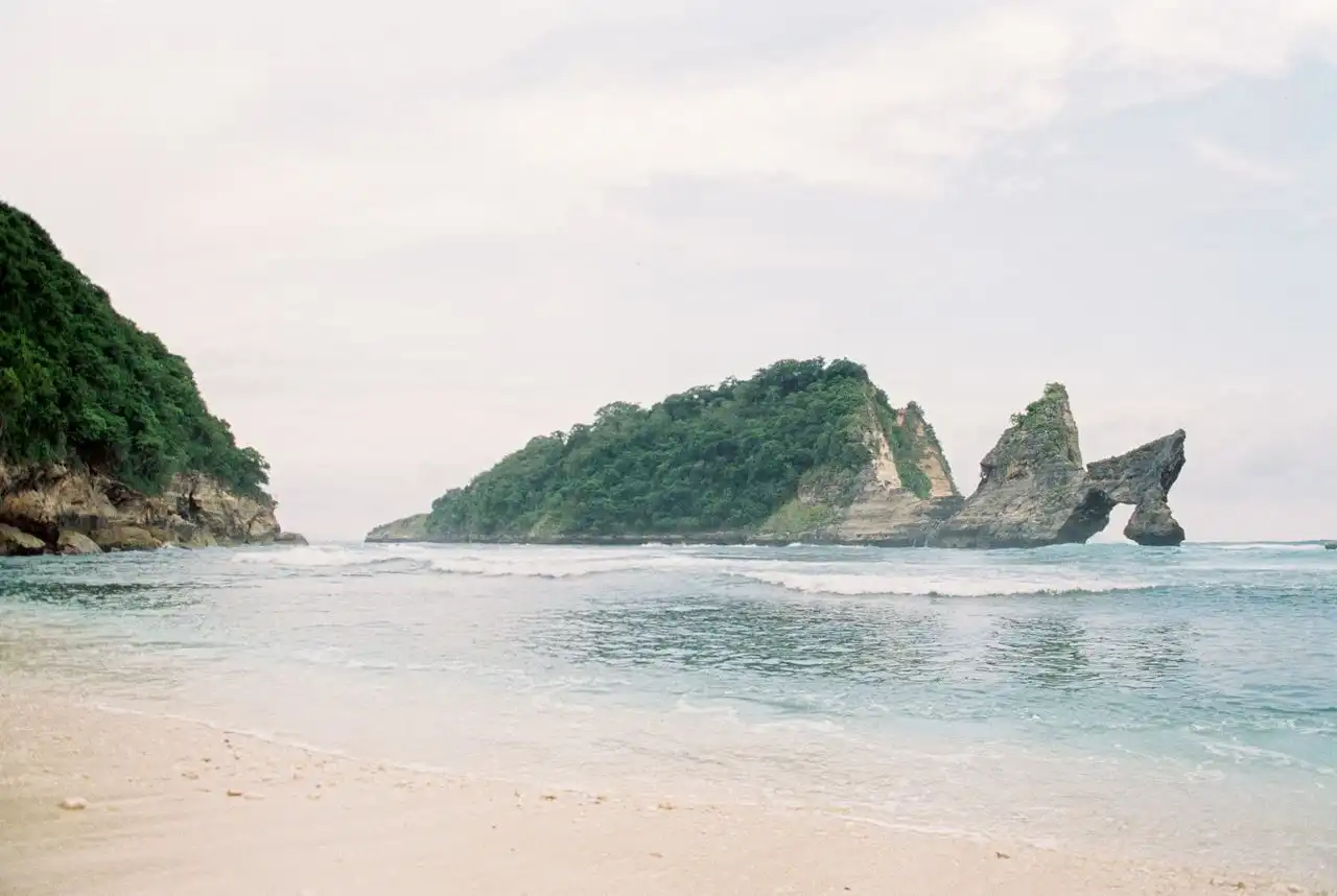 A calm white sand beach on Nusa Penida with a natural rock arch formation and a lush green rocky islet rising from the pale blue ocean, shot in a soft film photography aesthetic under an overcast sky