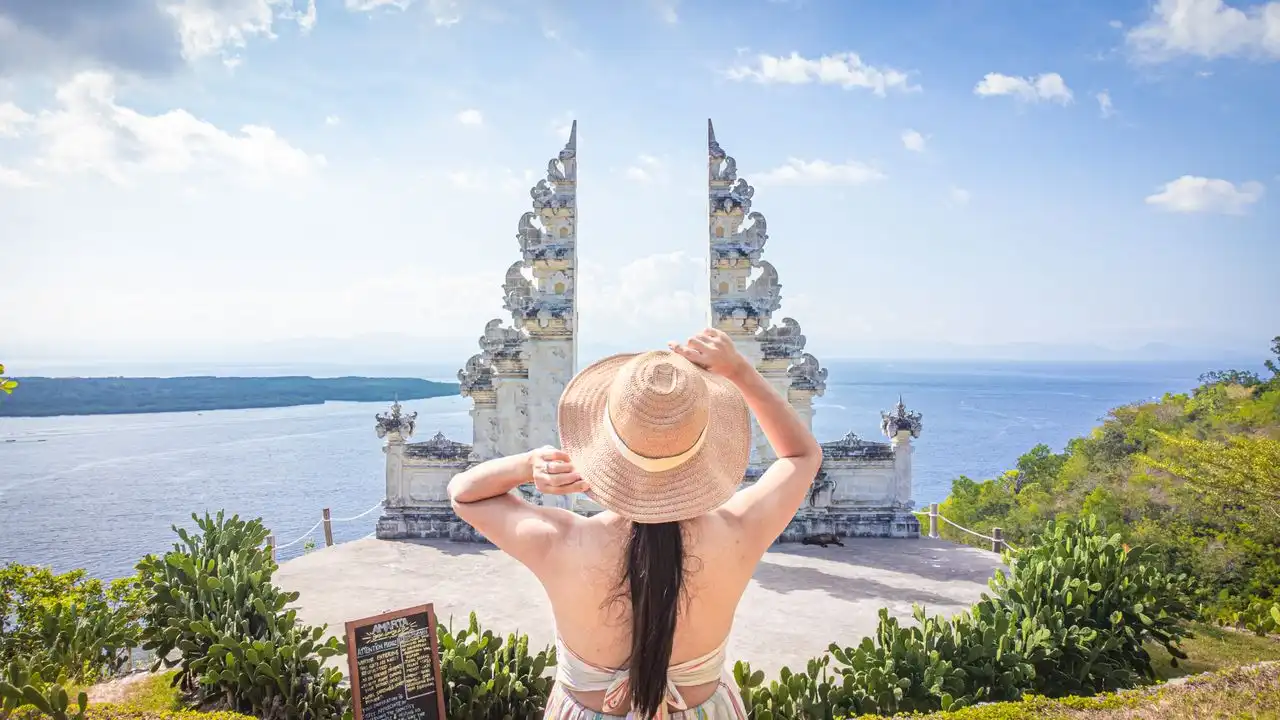 Behind-the-back view of a woman wearing a wide-brimmed straw hat, looking out through a traditional white Balinese split gateway (Candi Bentar). The gate overlooks a vast blue ocean under a bright, sunny sky with light clouds, with green hills visible in the distance.