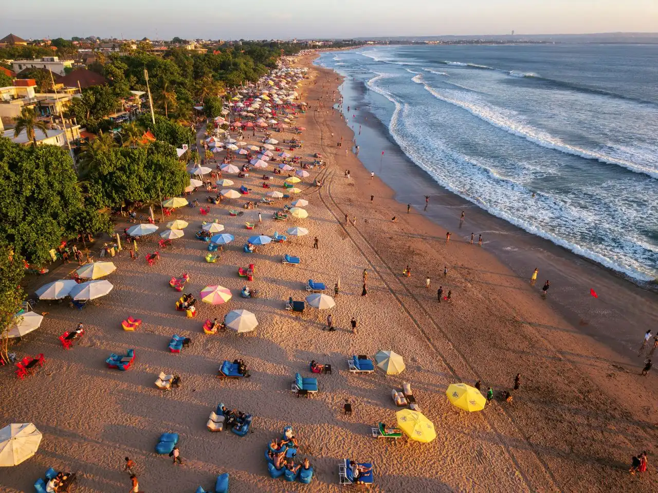 Aerial perspective of a crowded Balinese beach lined with colorful umbrellas and bean bags during a golden sunset, with waves crashing on the shore.