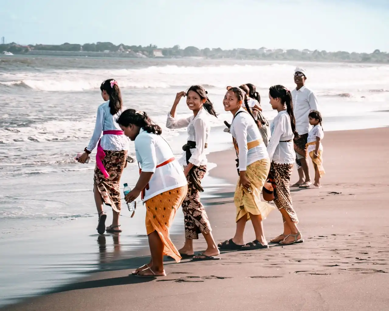 A group of Balinese women and children dressed in traditional ceremonial attire—white blouses (kebaya), colorful sashes, and patterned batik sarongs—walking along the shoreline of a sandy beach. Some turn to smile at the camera, with ocean waves crashing gently in the background.