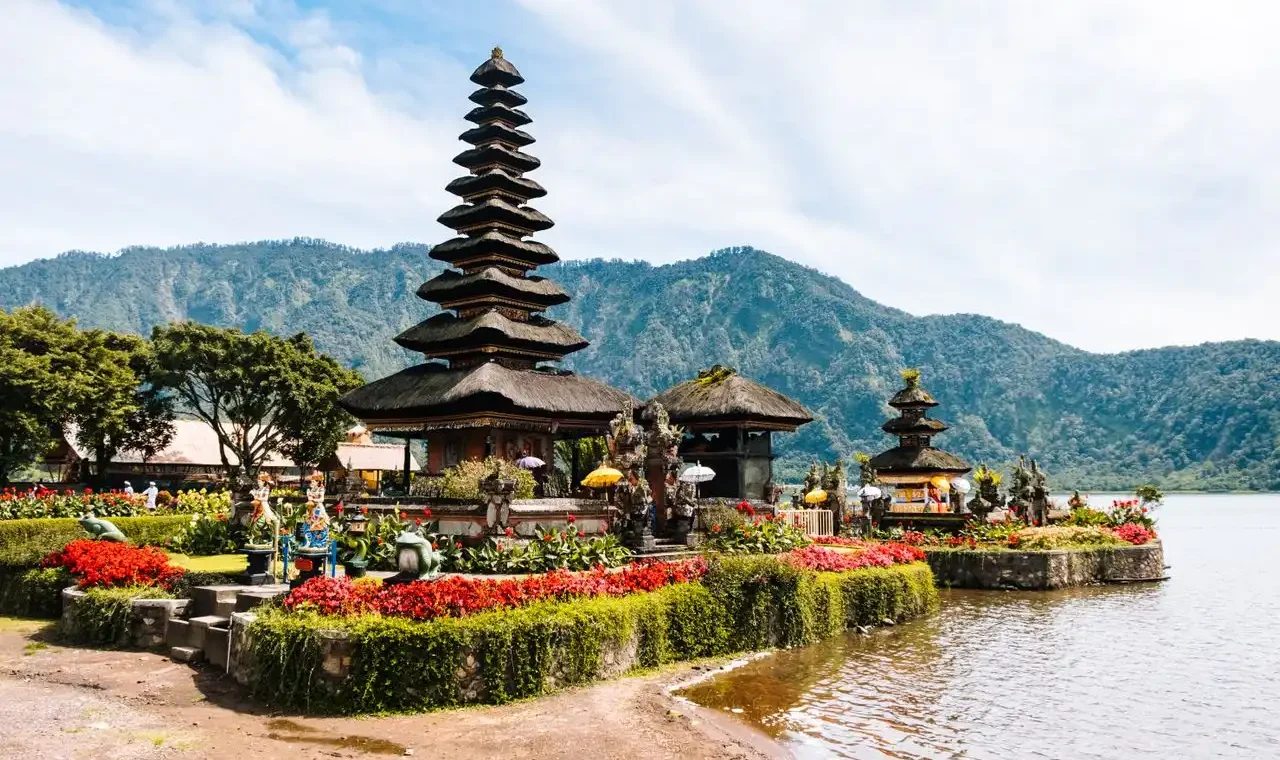 A scenic view of the iconic multi-tiered Meru tower of Ulun Danu Beratan Temple, situated on the edge of Lake Bratan in Bali, Indonesia. The temple is surrounded by manicured gardens with vibrant red flowers, with lush, mist-shrouded green mountains rising in the background under a blue sky