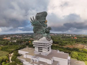 Aerial side view of the massive Garuda Wisnu Kencana Statue standing tall on a concrete pedestal surrounded by lush green landscape in Bali, Indonesia.