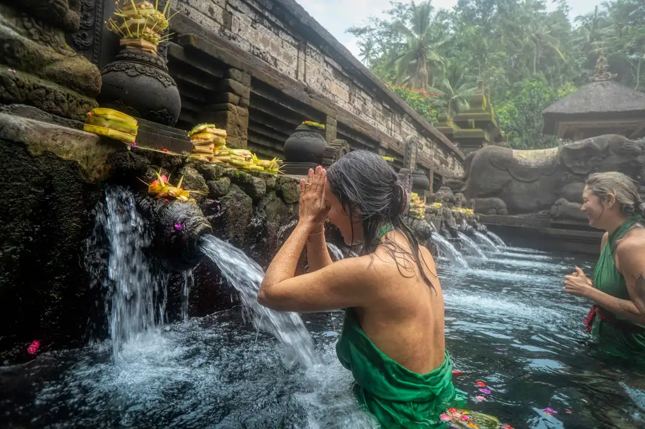 A close-up shot focusing on a woman in a green ritual robe, hands pressed together in prayer, with water pouring from a stone spout onto her head in a holy bathing pool at Pura Gunung Kawi Sebatu, Bali. Offerings sit on the ledge.