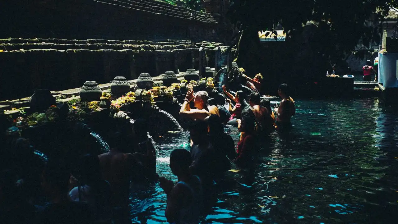 Devotees and visitors standing in line in the water pool, praying under stone water spouts decorated with offerings during a Melukat purification ceremony at Tirta Empul Temple in Bali.